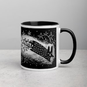 May include: A black and white mug with a black handle. The mug features a black and white illustration of a rabbit shooting across a starry sky. The background is black with white stars.
