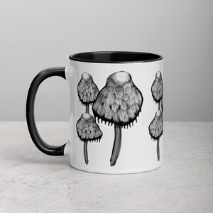 May include: White ceramic mug with a black rim and handle. The mug features a black and white illustration of mushrooms.