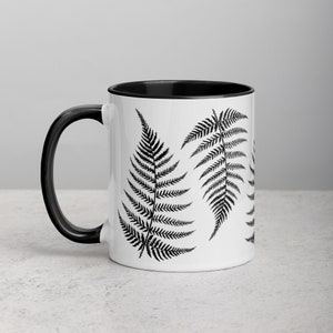 May include: A white ceramic mug with a black interior and handle. The mug features a black and white fern leaf design.