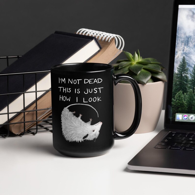May include: Black ceramic mug with the text "I'M NOT DEAD THIS IS JUST HOW I LOOK" and a white opossum illustration. The mug has a large handle and is displayed on a white surface.