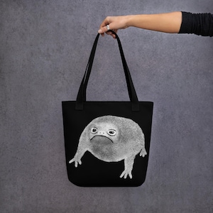 May include: Black tote bag featuring a white illustration of a frog with a grumpy expression.
