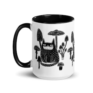 May include: White ceramic mug with a black interior and handle. The mug features a black and white illustration of a smiling cat surrounded by various mushrooms. The design is whimsical and artistic, suitable for coffee or tea.