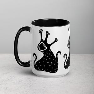 May include: A white ceramic coffee mug with a black rim and handle. The mug features a black and white illustration of a cartoon creature with multiple arms and a wide open mouth.