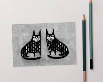Op de afbeelding: Een zwart-wit illustratie van twee lachende katten met verticale lijndetails. De katten staan op grijs papier, met twee potloden op de achtergrond, een groene en een blauwgroene.