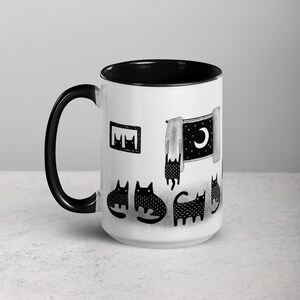May include: White ceramic coffee mug with a black rim and handle. The mug features a black and white illustration of cats looking at a window with a crescent moon.
