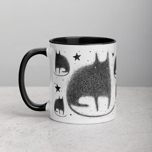 May include: White ceramic mug with a black rim and handle. The mug features a black and white illustration of a cat with a fluffy texture and stars. The cat is surrounded by smaller cats.