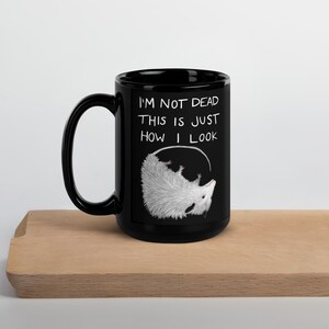 May include: Black ceramic mug with the text "I'M NOT DEAD THIS IS JUST HOW I LOOK" and a white illustration of an opossum. The mug has a large handle and is sitting on a wooden surface.