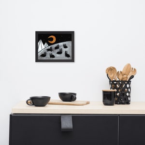 May include: A black framed print of a black and white illustration of a crescent moon, two white mountains, and a group of black rabbits against a dark blue background.
