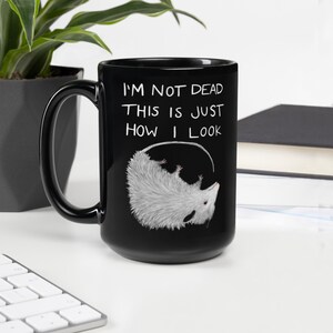 May include: Black ceramic mug with a white opossum illustration and the text "I'M NOT DEAD THIS IS JUST HOW I LOOK". The opossum is hanging upside down. The mug has a black handle.