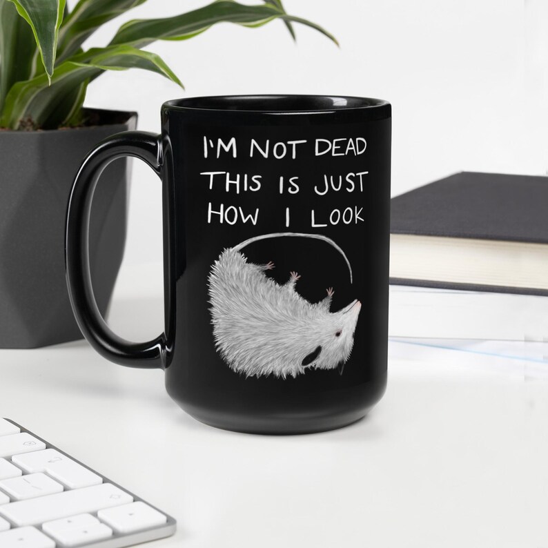May include: Black ceramic mug with a white opossum illustration and the text "I'M NOT DEAD THIS IS JUST HOW I LOOK". The opossum is hanging upside down. The mug has a black handle.