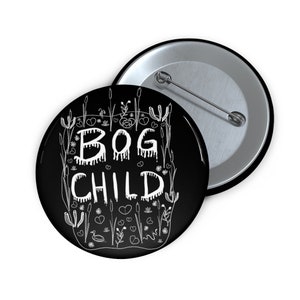 May include: A black button with a white design. The design features the words "BOG CHILD" in a dripping font, surrounded by stylized plants and flowers.