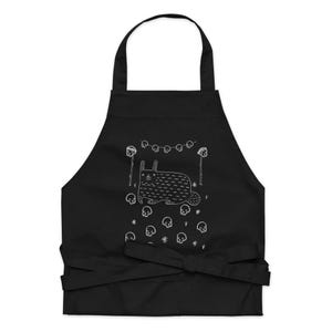 May include: Black apron with a white illustration of a cat surrounded by skulls and a string of lights. The cat is lying down and has a surprised expression.