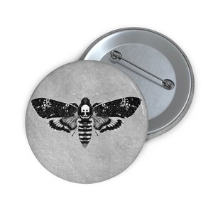 May include: A black and white button featuring a moth with a skull on its head. The moth is on a gray background.