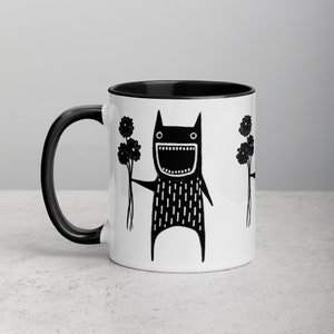 May include: A white ceramic mug with a black interior and handle. The mug features a black and white illustration of a cartoon monster holding a bouquet of flowers. The monster has a wide open mouth and a happy expression.