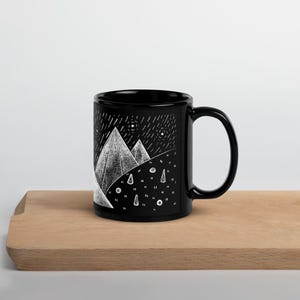 Pode incluir: Caneca de cer&acirc;mica preta com uma ilustra&ccedil;&atilde;o branca de uma cordilheira sob um c&eacute;u estrelado. O design inclui &aacute;rvores estilizadas e chuva. A caneca est&aacute; sobre uma superf&iacute;cie de madeira.