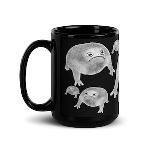 May include: Black ceramic mug with a white design of multiple frogs with grumpy expressions. The frogs are different sizes and are arranged in a repeating pattern.