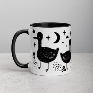 May include: Black and white coffee mug with a black rim. The mug features a design of two black ducks with a moon and stars in the background.