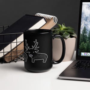 May include: Black ceramic mug featuring a white cartoon deer design. The deer has large antlers and a toothy grin. The mug is on a white surface with a laptop, a plant, and a wire basket with notebooks in the background.