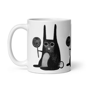 May include: White ceramic mug with a black and white illustration of a rabbit holding a lollipop. The rabbit has a skull-like face and is wearing a bow tie.