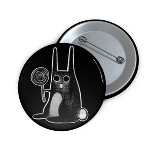 May include: A black button with a white outline of a rabbit holding a lollipop. The rabbit has large eyes and a toothy grin.