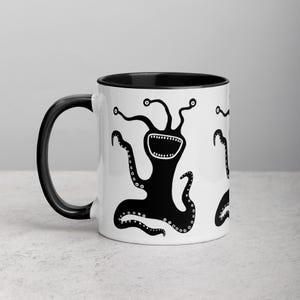 Könnte beinhalten: Weiße Keramik-Kaffeetasse mit schwarzem Rand und Henkel. Die Tasse zeigt eine Schwarzweißillustration eines dreäugigen, zahnigen Wesens mit Tentakeln.