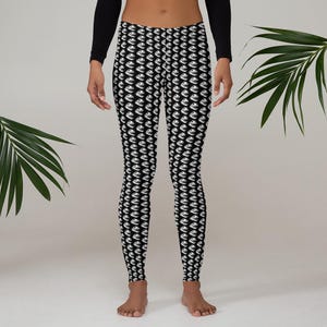 Peut inclure: Legging noir avec un motif blanc répété. Le legging est moulant et arrive aux chevilles. Le motif semble être un dessin stylisé. La personne portant le legging se tient devant un fond neutre.