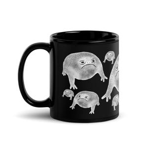 以下が含まれることがあります： 黒いセラミック製のコーヒーカップで、白いデザインで複数のカエルが描かれています。カエルは不機嫌そうな表情をしています。カエルは様々な大きさで、様々な位置に描かれています。