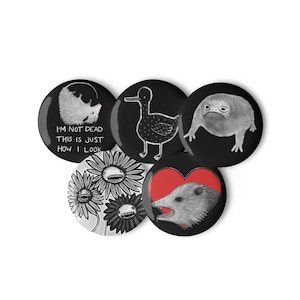May include: A set of five circular pin-back buttons featuring black and white illustrations. One button shows an opossum with the text "I'M NOT DEAD THIS IS JUST HOW I LOOK," a duck, a toad, three flowers with faces, and an opossum inside a red heart.