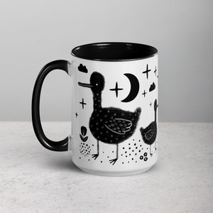 Könnte beinhalten: Eine weiße Keramiktasse mit schwarzem Rand und Henkel. Die Tasse zeigt eine Schwarzweißillustration von zwei Enten mit einem Halbmond und Sternen im Hintergrund.