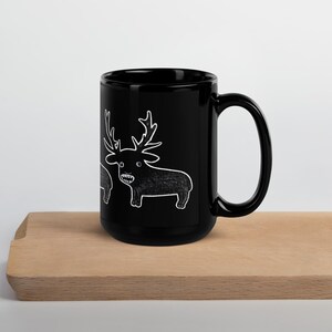 May include: A black ceramic mug with a white cartoon deer illustration. The deer has large antlers and a toothy grin. The mug is sitting on a wooden surface.