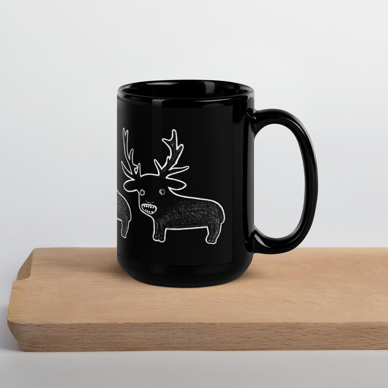 May include: A black ceramic mug with a white cartoon deer illustration. The deer has large antlers and a toothy grin. The mug is sitting on a wooden surface.