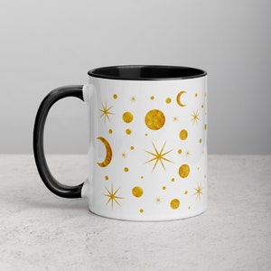 May include: White ceramic mug with a black rim and handle. The mug features a gold pattern of moons, stars, and dots on a white background.
