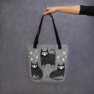 Op de afbeelding: Een grijze tote bag met zwarte banden en een zwart-wit ontwerp van drie katten met schedelgezichten. De katten zijn omringd door witte sterren.