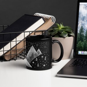 Pode incluir: Caneca de cer&acirc;mica preta com uma ilustra&ccedil;&atilde;o de montanha e chuva branca. A caneca tem uma al&ccedil;a preta e um acabamento brilhante. Uma cesta de arame com cadernos e um laptop com uma cena de montanha est&atilde;o no fundo.