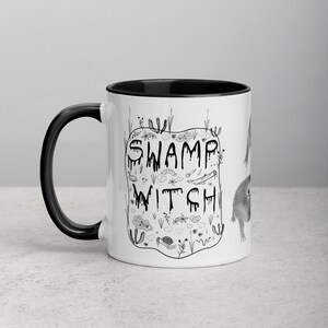 May include: White ceramic mug with a black rim and handle. The mug features a black and white illustration of a swamp witch with the text "Swamp Witch" in a dripping font.