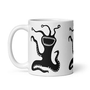 May include: White ceramic mug featuring a black cartoon monster design. The monster has multiple tentacles, large eyes, and a wide open mouth. The mug has a curved handle and a glossy finish.