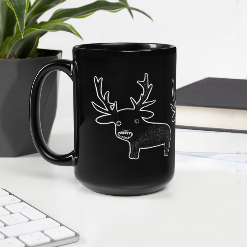 May include: Black ceramic mug featuring a whimsical cartoon deer illustration. The deer is drawn in white with large antlers, a small body, and a toothy grin. The mug has a large handle and is set against a neutral background.