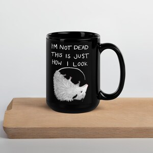 May include: Black ceramic mug with a white opossum illustration and the text "I'M NOT DEAD THIS IS JUST HOW I LOOK". The opossum is drawn upside down inside a white circle.