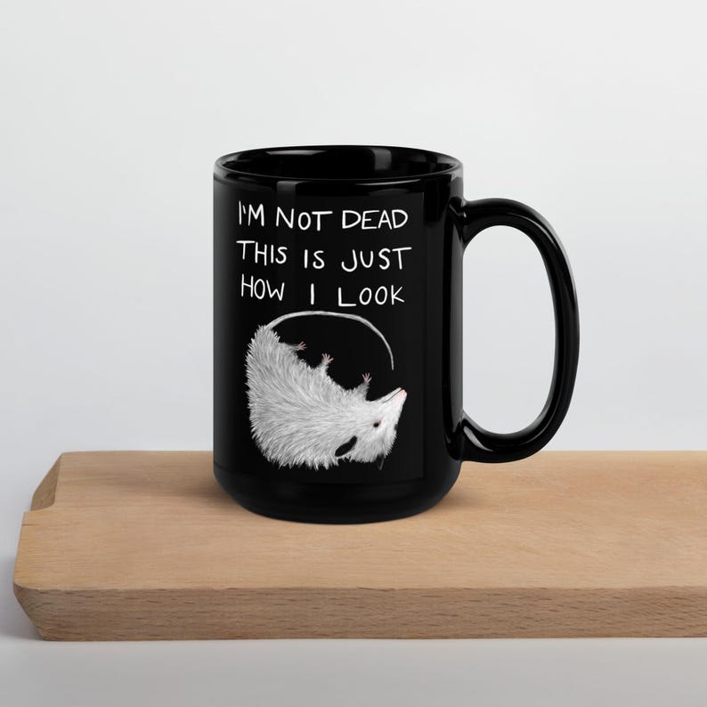 May include: Black ceramic mug with a white opossum illustration and the text "I'M NOT DEAD THIS IS JUST HOW I LOOK". The opossum is drawn upside down inside a white circle.