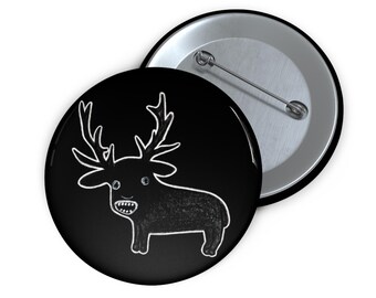 May include: A black button featuring a white outline of a cartoon deer with large antlers. The deer has a wide, toothy grin.