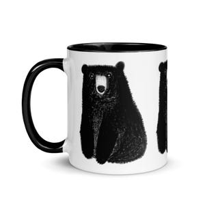 May include: White ceramic coffee mug with a black rim and handle. The mug features a black and white illustration of a bear on the front.
