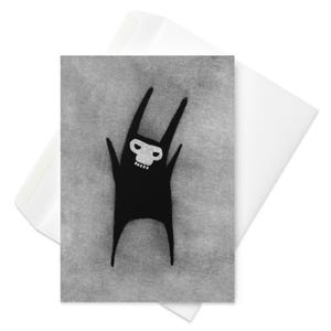 May include: Greeting card featuring a black creature with a white skull-like face and long ears, arms raised in a celebratory pose. The card is set against a textured gray background, with a white envelope partially visible.