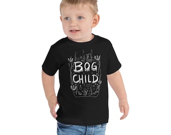 Op de afbeelding: Een zwart peuter T-shirt met de witte tekst "BOG CHILD", omringd door een grillig ontwerp. Het kind draagt een blauwe spijkerbroek.