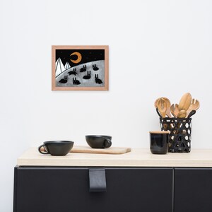 May include: A framed illustration of a black and white landscape with a crescent moon and a group of black rabbits. The moon is a bright orange color. The background is a dark gray color. The rabbits are all facing the same direction. The illustration is in a wooden frame.