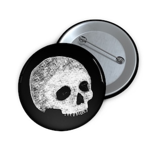 May include: A black and white button featuring a white skull with a textured, chalk-like appearance. The skull is facing to the right and has a detailed, realistic design.
