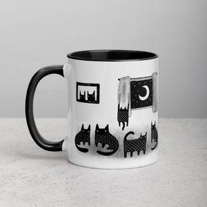 May include: White ceramic mug with a black rim and handle. The mug features a black and white illustration of cats looking out a window with a moon and stars in the sky.