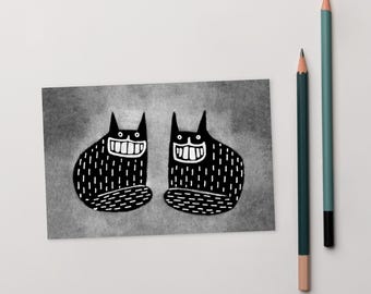 Op de afbeelding: Twee zwarte katten met witte ogen en tanden zitten naast elkaar op een grijze achtergrond. Ze lachen allebei met hun mond open.