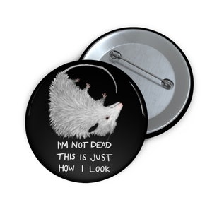 May include: A black and white button with a possum on it. The possum is curled up in a ball with its tail curled around its body. The text "I'm not dead this is just how I look" is printed below the possum.