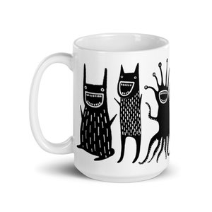 May include: White ceramic mug with a black and white illustration of three cartoon monsters with big smiles. The monsters are all different shapes and sizes, and they are all facing forward. The mug is a standard size and has a handle on the right side.