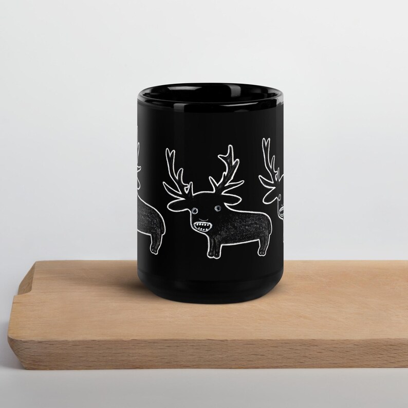 May include: Black ceramic mug featuring a repeating white cartoon deer design. The deer have large antlers and are smiling. The mug is sitting on a light brown wooden surface.